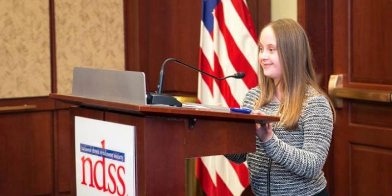 Girl with Down syndrome speaks at a podium with an NDSS sign in front of a United States flag