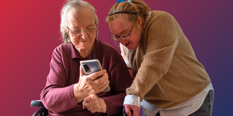 A woman with Down syndrome looks at a smart phone with an older woman sitting in a mobility device