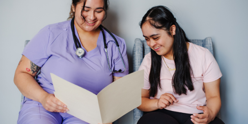 A young woman with Down syndrome looks at a file that a healthcare professional wearing lilac scrubs shares with her